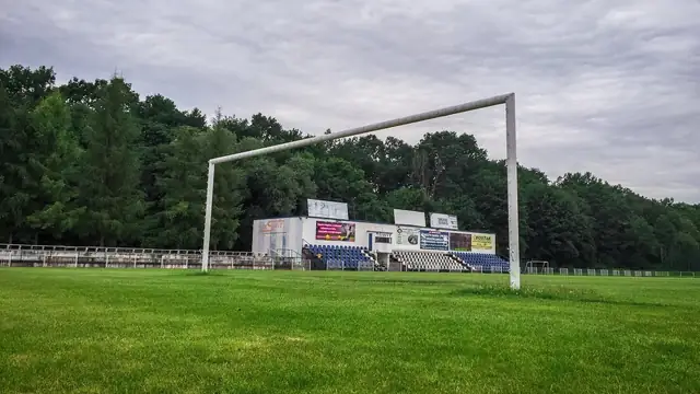 A soccer goal stands on an empty grass field in front of a small grandstand and trees