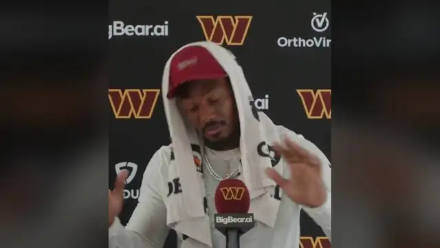 Stefon Diggs in a red cap with a towel over his head speaking at a Commanders press conference podium