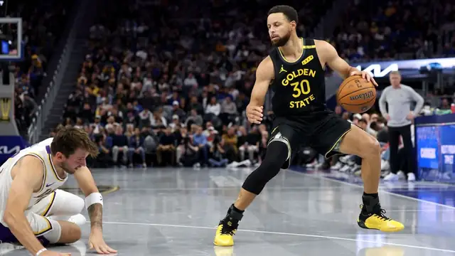 Stephen Curry in a black Golden State number 30 jersey dribbles as Luka Doncic of the Lakers falls to the floor at Chase Center