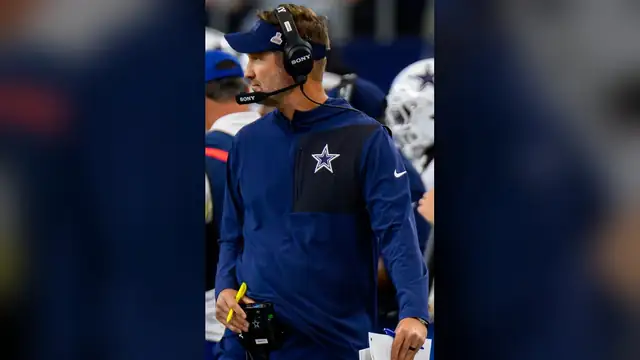 Brian Schottenheimer in a navy Cowboys quarter-zip and headset walks the sideline holding a play sheet at AT&T Stadium in October 2025