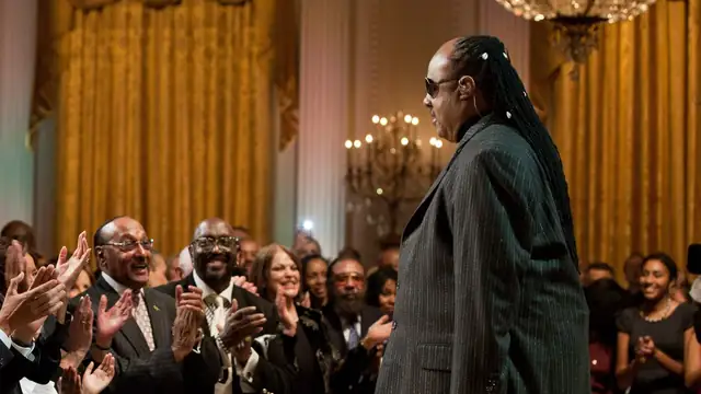 Stevie Wonder in dark glasses and a pinstriped suit faces a crowd giving him a standing ovation in the gold-curtained East Room of the White House