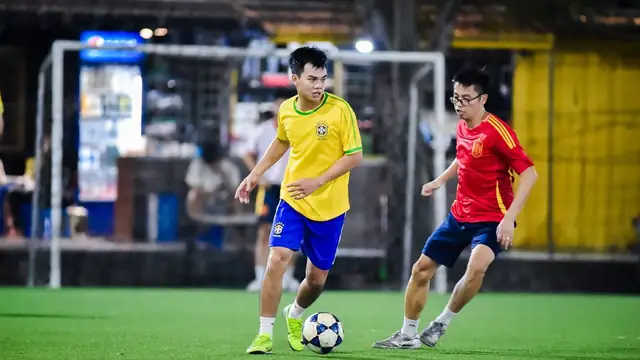 Two young men chasing a soccer ball on an artificial turf field lit up at night in front of a goal