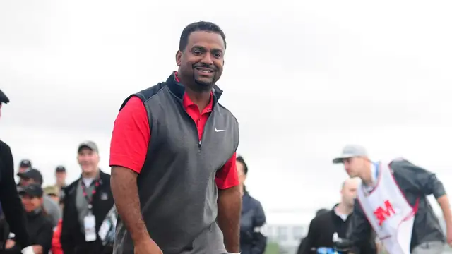 Alfonso Ribeiro grins in a red polo and gray vest as he walks across a golf course in front of a crowd