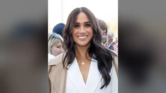 Meghan Markle smiling with long dark wavy hair in a white blazer and camel coat at an outdoor event