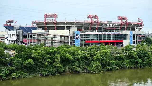 Nissan Stadium in Nashville with its red light towers and a Titans banner on the facade rises above green trees along the Cumberland River