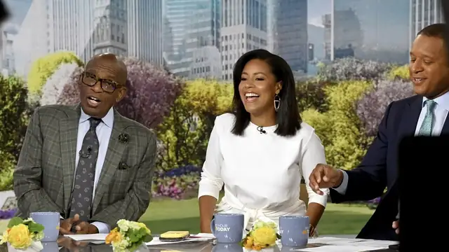 Morning show hosts laughing together at a studio desk with coffee mugs and yellow flowers in front of a city skyline backdrop