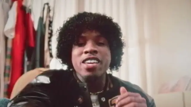 Tory Lanez with curly hair in a black varsity jacket talking in a room beside a clothing rack