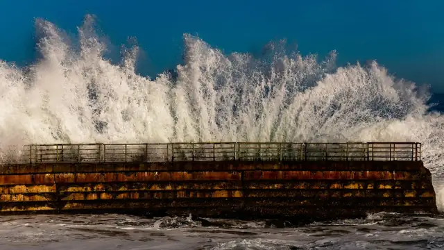 A large ocean wave crashes over a concrete pier as white spray rises against a cloudy sky