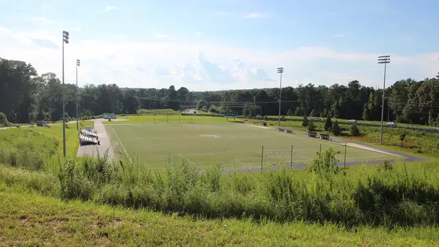 An empty artificial turf soccer field ringed by light poles and a paved walkway with tall grass and trees under a summer sky