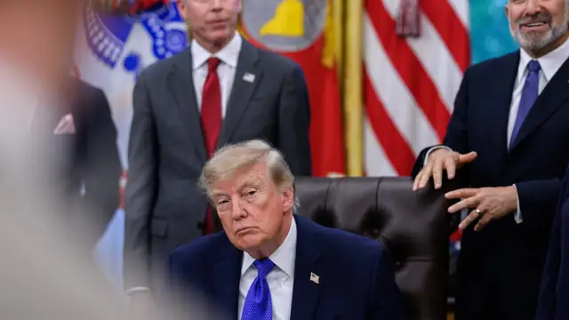 President Donald Trump sits at his desk in the Oval Office as two officials stand behind him in front of flags