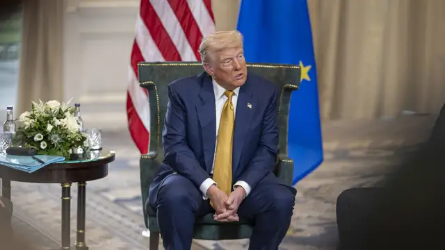 Donald Trump in a navy suit and gold tie seated in a green armchair before American and European Union flags