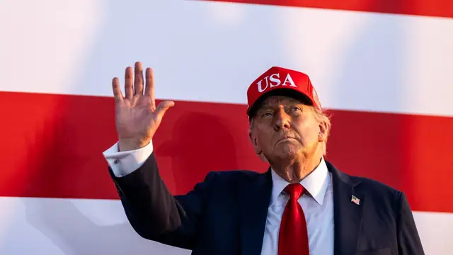 President Donald Trump in a red USA cap waves to supporters in front of a giant American flag at an outdoor rally
