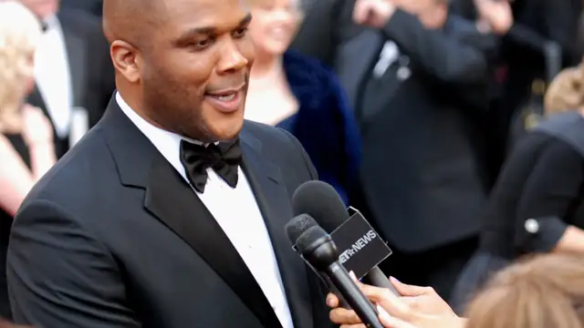 Tyler Perry in a black tuxedo speaking into a microphone for reporters at a crowded red carpet event