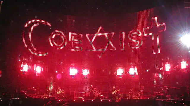 U2 performs on a dark stage under a giant red lighted Coexist sign during a Vertigo Tour concert