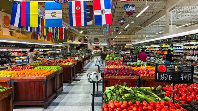 A grocery store produce section with fruit displays and flags of Latin American countries hanging from the ceiling