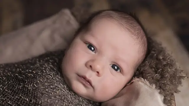 A newborn baby with big blue eyes lies wrapped in a soft brown knit blanket looking up at the camera