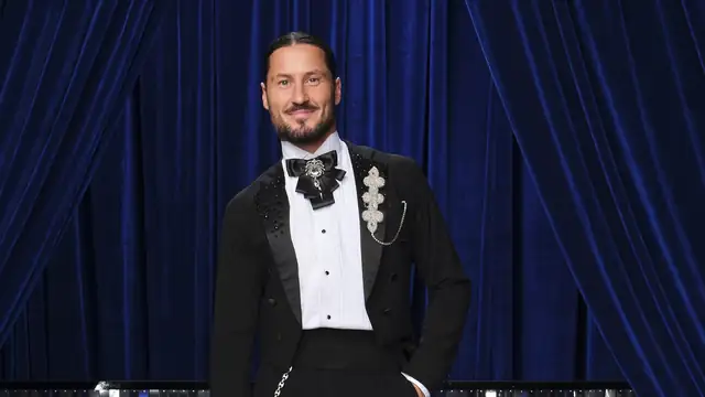 Val Chmerkovskiy in a black tailcoat with a bow tie posing in front of a blue velvet curtain