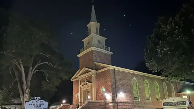 The steepled brick First Baptist Church in Williamsburg lit at night with a historical marker in the front yard