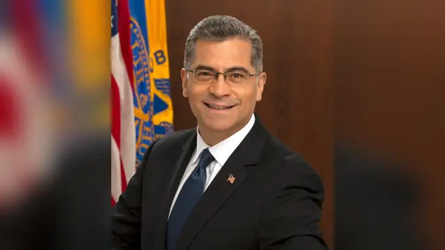 Xavier Becerra smiling in a dark suit and blue tie in his official portrait beside an American flag
