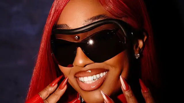 Yung Miami smiling in red hair and dark wraparound sunglasses