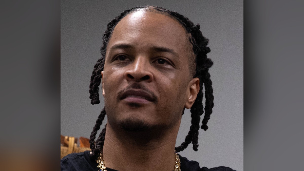 Rapper T.I. with long locs in a black T-shirt and a gold gemstone chain looking off to one side against a gray wall