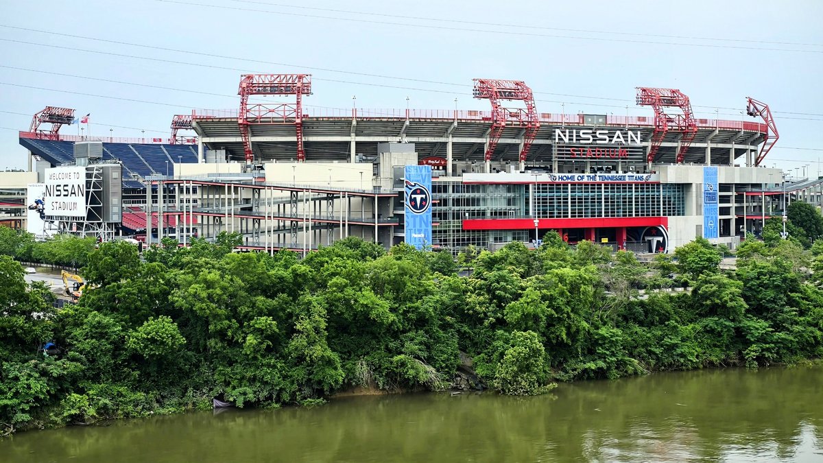 Nissan Stadium in Nashville with its red light towers and a Titans banner on the facade rises above green trees along the Cumberland River