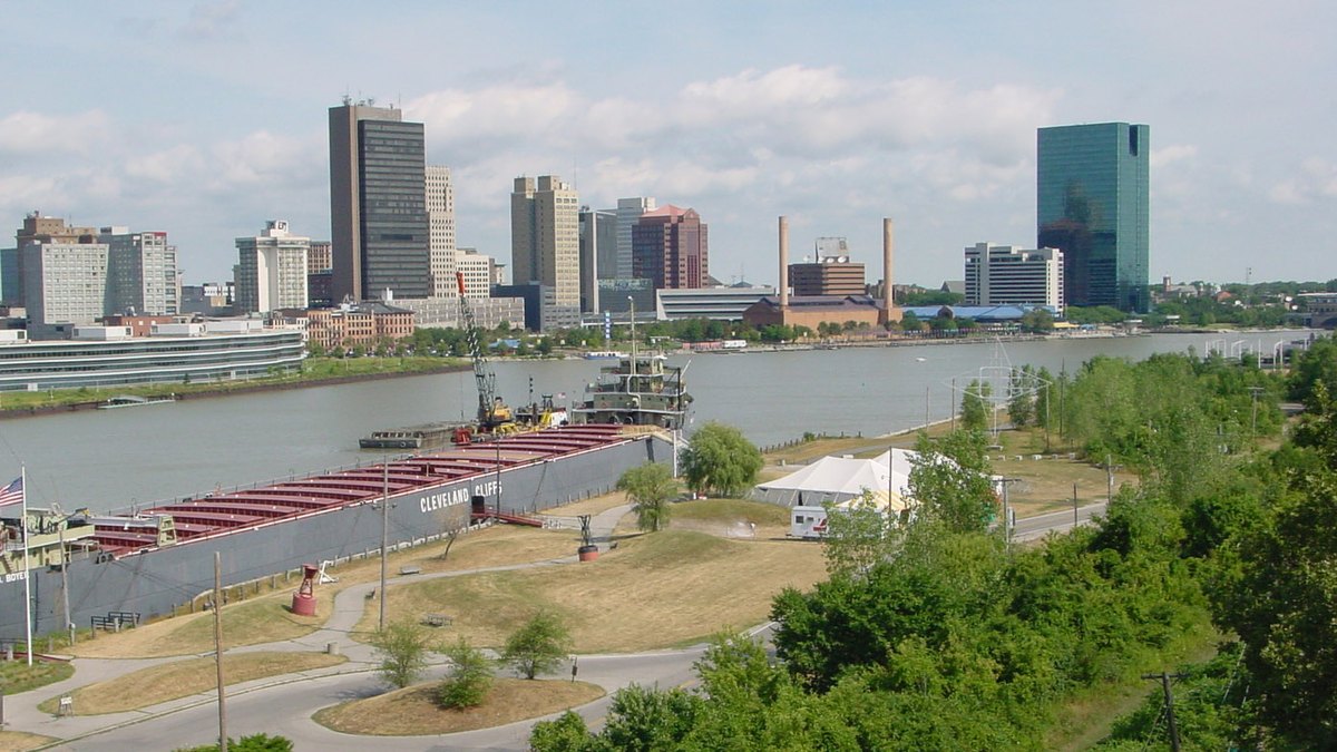 Downtown Toledo office towers rising beyond the Maumee River on a summer day with a retired freighter docked along a grassy riverside park
