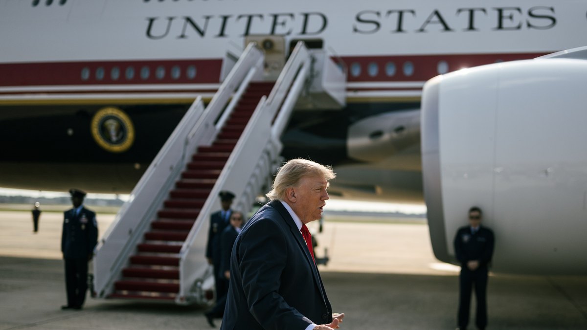 President Donald Trump in a navy suit and red tie talks to reporters on the tarmac in front of Air Force One