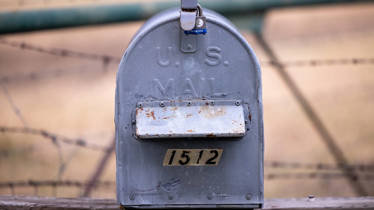 A weathered rural U.S. mailbox on a post beside a barbed-wire fence