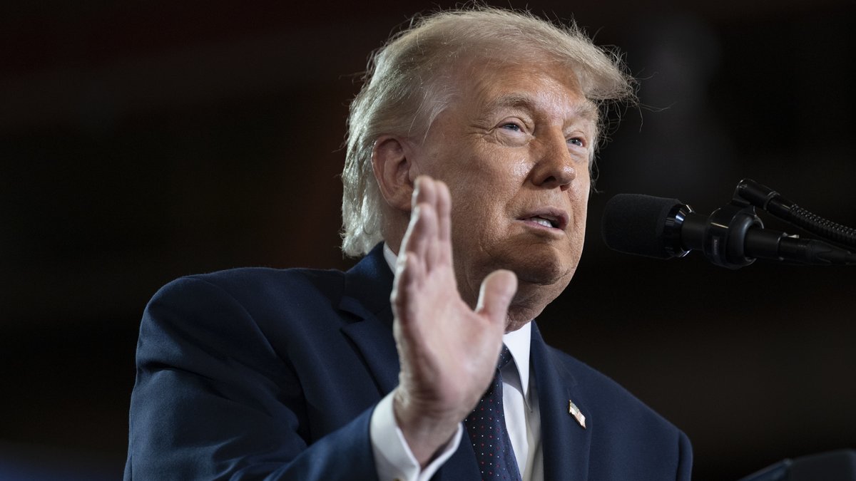 President Donald Trump raising his hand as he speaks into a microphone in a navy suit