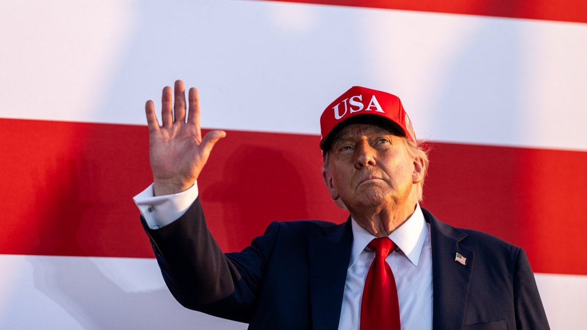 President Donald Trump in a red USA cap waves to supporters in front of a giant American flag at an outdoor rally