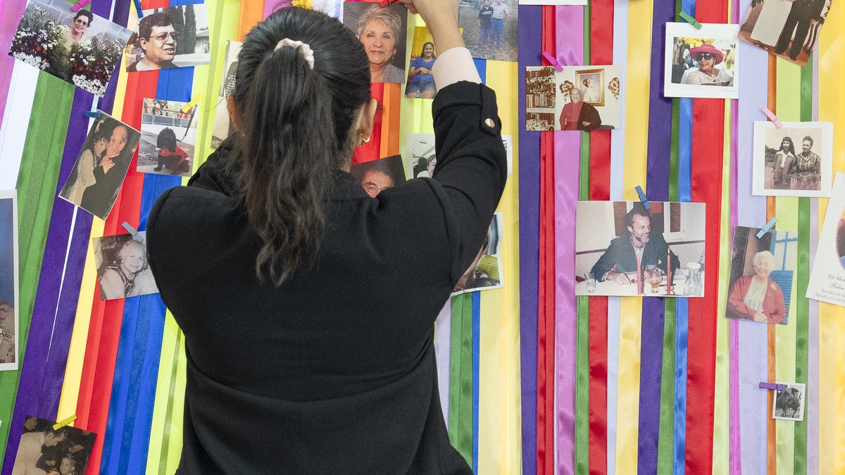 A woman pins a family photo onto a colorful ribbon ofrenda decorated with pictures of loved ones