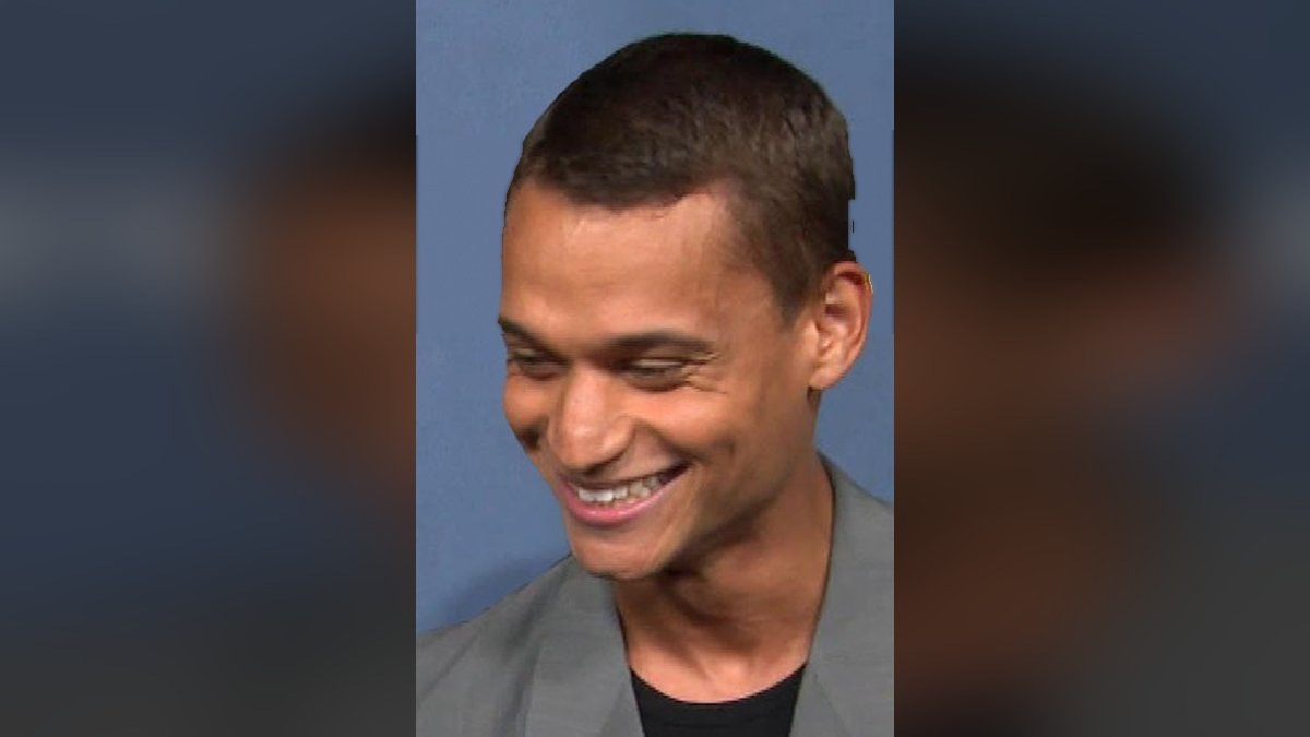 Tyriq Withers laughing in a gray blazer in front of a blue event backdrop