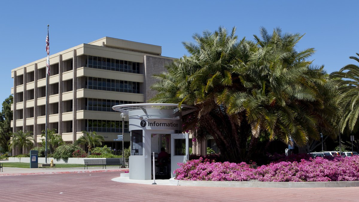 A multistory campus office building behind a small entrance kiosk with palm trees and pink flower beds at the University of California, Santa Barbara