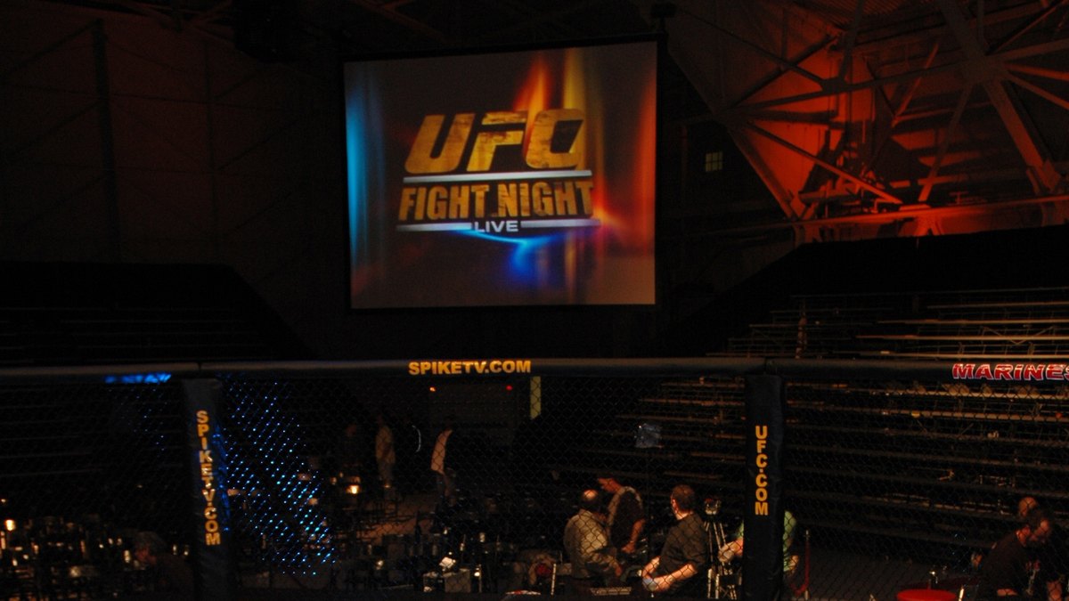 An empty UFC Octagon lit in blue under a big screen before a UFC Fight Night event