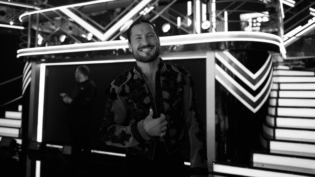 Val Chmerkovskiy smiling with a thumbs-up in a black-and-white photo on the Dancing with the Stars stage