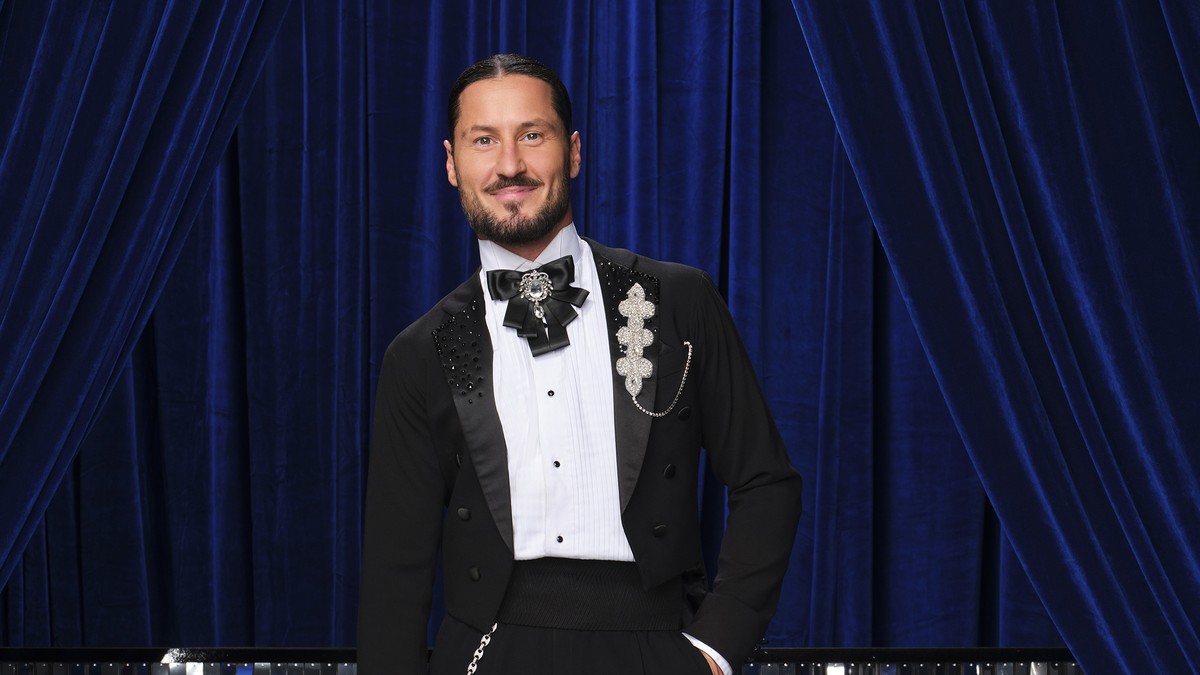Val Chmerkovskiy in a black tailcoat with a bow tie posing in front of a blue velvet curtain