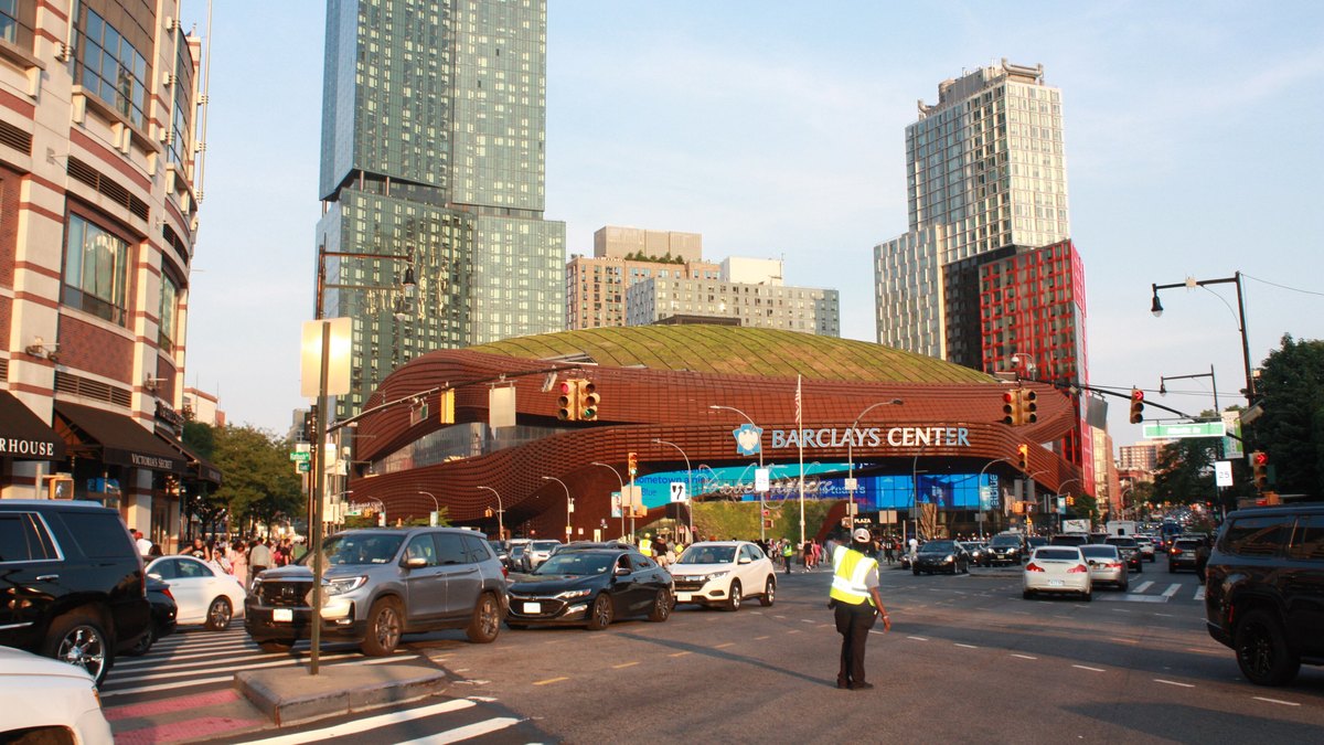 Barclays Center arena in Brooklyn with its rust-colored steel canopy and city towers behind it on a sunny day