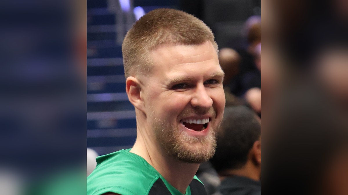 Kristaps Porziņģis smiling in a green and black Celtics warmup shirt inside an arena