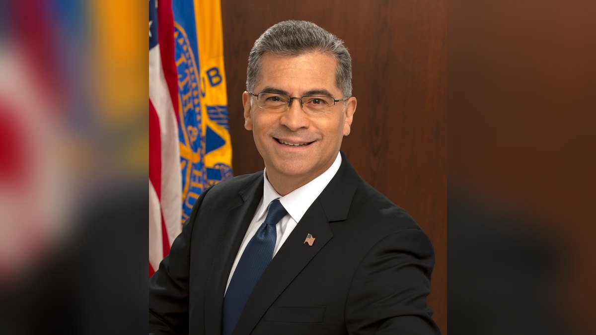 Xavier Becerra smiling in a dark suit and blue tie in his official portrait beside an American flag