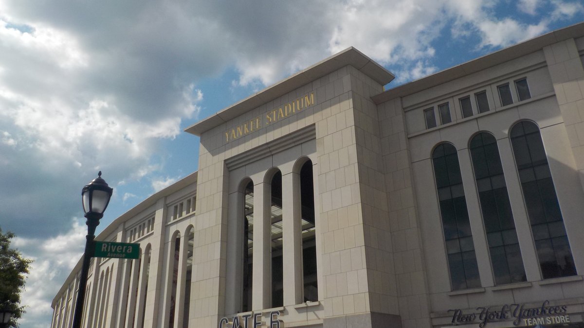The limestone front of Yankee Stadium with its arched windows and gold Yankee Stadium lettering under a cloudy summer sky in the Bronx
