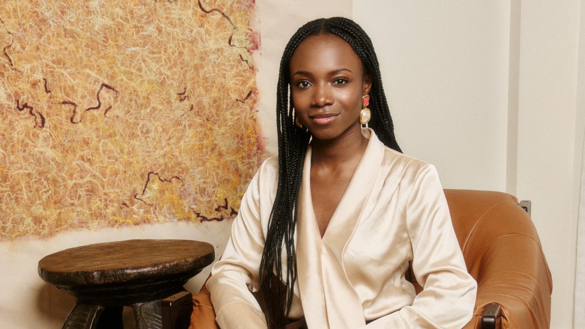 Yinka Ogunbiyi in long box braids and a cream satin blouse smiling in a leather armchair beside a woven wall hanging